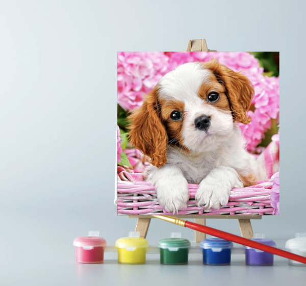 An easel with paints and a puppy in a basket paint by number canvas on it